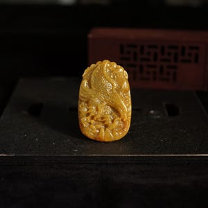 以下が含まれることがあります： 翼を広げた鳥の複雑なレリーフ彫刻が施された、楕円形のゴールデンイエローのペンダント。ペンダントの表面にはデザイン全体に詳細な質感と模様が見られ、磨き上げられた半透明の仕上がりとなっています。