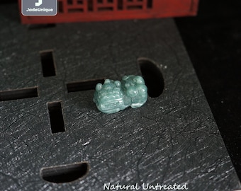 Puede incluir: Pequeña figura tallada de Pixiu en jadeíta verde. Se encuentra sobre una superficie gris oscuro con el texto "Natural Untreated Guatemalan Jadeite Pixiu". El logotipo de JadeUnique está en la esquina superior izquierda.