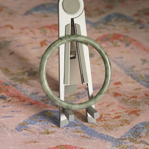 May include: A smooth, circular jade bangle with a pale green hue and subtle darker green inclusions. The bangle is displayed upright on a silver-colored, adjustable metal stand with a central hinge and two support arms at the base.