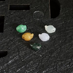 Könnte beinhalten: Eine Sammlung kleiner, geschnitzter Schildkrötenfiguren aus Jade in verschiedenen Farben, darunter Grün, Gelb, Weiß und Dunkelgrün. Die Schildkröten sind auf einer strukturierten schwarzen Oberfläche ausgestellt.