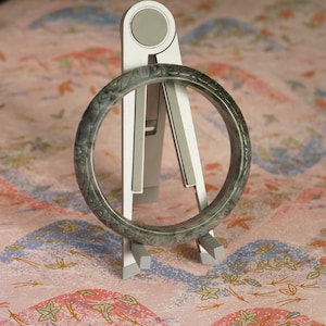 Puede incluir: Un brazalete de piedra gris oscuro y verde translúcido con intrincados patrones florales tallados en su superficie exterior, exhibido verticalmente sobre un soporte metálico plegable plateado con una parte superior circular.