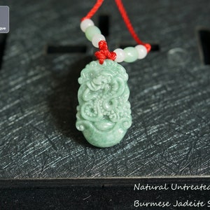 May include: A green jadeite snake pendant with intricate carvings. The pendant is on a red string with white beads. The text "Natural Untreated Burmese Jadeite Snake" is below the pendant.