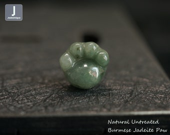 Puede incluir: Pequeña talla pulida en forma de pata de jadeíta birmana verde. La piedra preciosa natural sin tratar presenta almohadillas redondeadas e incisiones de garras. El texto "Natural Untreated Burmese Jadeite Paw" es visible.