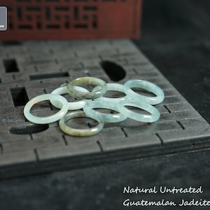 May include: A group of six natural, untreated Guatemalan jadeite rings in various shades of green and white. The rings are stacked on a dark grey textured surface.