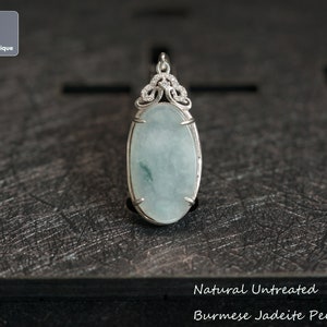 May include: A natural, untreated jadeite pendant from Burma. The pendant is oval shaped and set in silver with a delicate, ornate design. The jadeite is a light green color with a translucent quality.