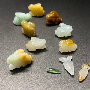 May include: A collection of ten jade rabbit figurines, some with brown and white coloring, and a jade carrot charm. The figurines are carved in a variety of poses, and the carrot charm is a simple, elongated shape.