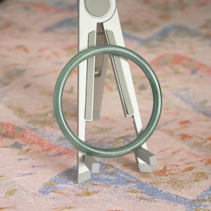 May include: A smooth, translucent, pale green circular bangle bracelet resting on a silver-colored metal stand. The bangle has a consistent, rounded profile and a polished finish, displaying a soft, uniform light green hue throughout its entire structure.