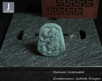 Peut inclure: Un pendentif en jadeïte vert en forme de dragon, sculpté dans un design détaillé. Le pendentif est naturel et non traité, et est fait de jadeïte du Guatemala. Le pendentif est sur une surface brun foncé avec un motif décoratif.