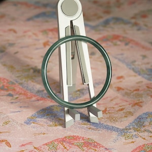 May include: A smooth, circular dark green jade bangle bracelet is displayed upright on a silver-toned, foldable metal stand. The bangle has a consistent, polished finish and a deep, uniform forest green color throughout its entire rounded structure.