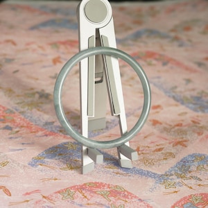 May include: A smooth, translucent, pale green circular bangle bracelet is displayed upright on a silver and gray folding stand. The bangle has a consistent, rounded profile and a polished finish, showcasing a subtle, uniform light green hue throughout.