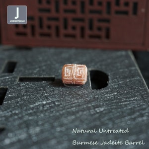 May include: Natural untreated Burmese jadeite barrel bead with a carved geometric pattern. The bead is a light brown colour and is sitting on a dark brown surface.