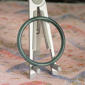 May include: A smooth, circular dark green bangle bracelet with a polished finish. The bangle is displayed upright on a silver-colored metal stand that features two vertical supports and two small horizontal prongs at the base for stability.
