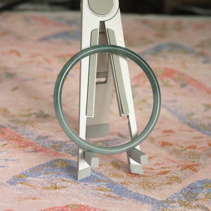 May include: A smooth, circular bangle bracelet with a translucent, pale green hue. The bangle is displayed resting on a silver-colored, foldable stand that features two vertical arms and a central support structure with grey rubberized grip pads.