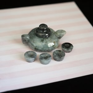 May include: A miniature teapot with a matching lid and four small cups, all carved from a polished, mottled grey and green stone. The teapot features a rounded body, a short spout, and a handle, while the four cups are small and circular.