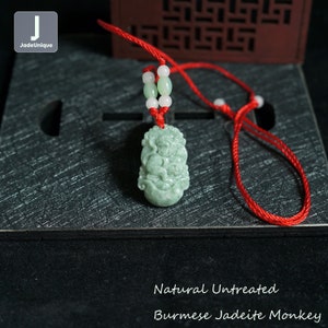 May include: A jadeite monkey pendant carved from natural, untreated Burmese jade. The pendant is green and has a red string attached to it. The pendant is on a black surface with a red string.