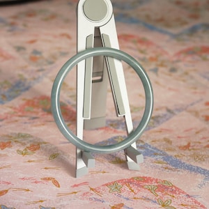 May include: A smooth, pale green circular bangle bracelet rests on a silver-colored, foldable metal stand. The bangle has a consistent, rounded profile and a soft, matte finish, positioned upright against the support structure of the stand.