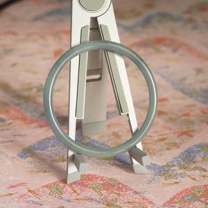 May include: A smooth, circular, sage green bangle bracelet is displayed upright on a silver-toned metal stand. The bangle has a solid, matte finish and a consistent, rounded profile, showcasing a simple and clean design without any additional ornamentation.