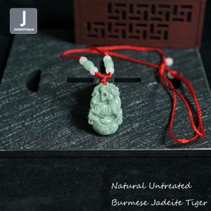 Pode incluir: Um pingente de jadeíta verde em forma de tigre em um colar de cordão vermelho. O pingente é esculpido com detalhes intrincados e tem um acabamento natural não tratado. O colar é feito com pequenas contas brancas.