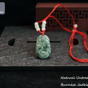May include: A green jadeite ox pendant carved with intricate details, hanging from a red string necklace with white beads. The pendant is on a dark gray surface. Natural Untreated Burmese Jadeite Ox.