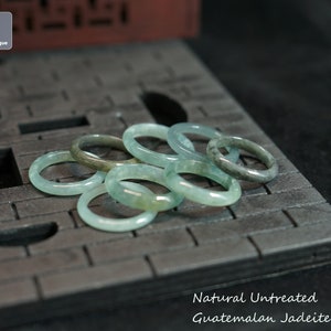 Peut inclure: Une collection d'anneaux en jadéite lisses et circulaires, dans des tons variés de vert pâle translucide et de gris tacheté. Les anneaux sont disposés en groupe. Le texte indique : J JadeUnique, Natural Untreated Guatemalan Jadeite Ring.