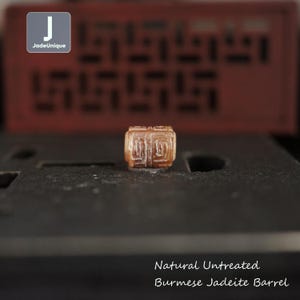 Puede incluir: Una cuenta de barril de jadeíta birmana natural sin tratar. La cuenta es de color marrón claro con patrones geométricos tallados. El texto "Natural Untreated Burmese Jadeite Barrel" está en la parte inferior de la imagen.