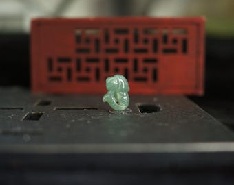 Puede incluir: Una pequeña talla de jade verde claro translúcido con forma de pez o criatura acuática estilizada. La talla presenta una superficie lisa y pulida con un pequeño orificio circular cerca de la base, mostrando variaciones de color naturales.