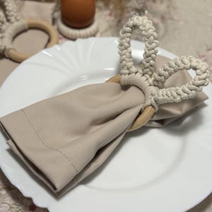 Puede incluir: Un plato blanco con una servilleta beige doblada sujeta por un servilletero de macramé. Un huevo marrón descansa sobre un pequeño posavasos tejido. También se ven una copa de vino y cubiertos.