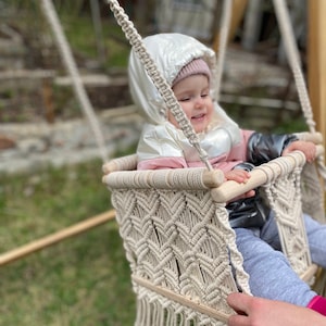 Op de afbeelding: Een baby zit in een macramé schommel met een houten frame. De schommel heeft een franje rand en een ruitpatroon. De baby draagt een roze en witte jas met een witte capuchon.