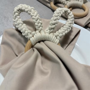 Puede incluir: Anillos de servilleta de macramé color crema con anillos de madera. Los anillos de servilleta están en bucle y anudados, creando un diseño texturizado. Las servilletas son de color beige claro, dobladas y colocadas sobre un plato blanco. Tema: decoración del hogar.