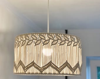 May include: A cream-colored macrame pendant light fixture. The lampshade has a rectangular shape with intricate woven patterns, including leaf and zigzag designs. The light hangs from a white ceiling fixture.