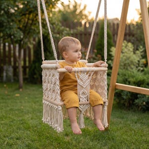 May include: A baby sits in a beige macrame swing with wooden accents, suspended from a wooden frame. The swing has a woven design and fringe detailing. The baby wears a mustard-colored outfit and is in a grassy outdoor setting.