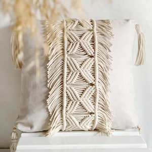 May include: A cream-colored pillow with a woven macrame design and fringe trim. The pillow is sitting on a white stool.