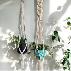 Puede incluir: Dos colgadores de plantas de macramé con plantas. Uno es blanco con una maceta negra, el otro es beige con una maceta turquesa. Ambos cuelgan contra una pared blanca con plantas verdes.