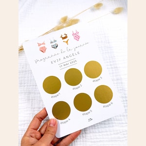 Könnte beinhalten: Eine weiße Karte mit dem Text "Programme de la journée EVJF ANGELE 17 MAI 2025" und sechs goldenen Rubbelkreisen. Die Karte zeigt Aquarellillustrationen von Badeanzügen. Von einer Hand gehalten.
