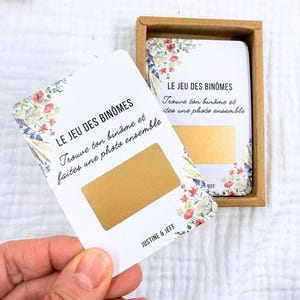 Könnte beinhalten: Ein Set weißer Karten mit floralen Akzenten und goldenen Rubbelbereichen. Auf den Karten steht "LE JEU DES BINÔMES" und "Trouve ton binôme et faites une photo ensemble." Die Karten befinden sich in einer braunen Box, mit den Namen "JUSTINE & JEFF".