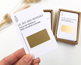 May include: White cards with gold rectangles and French text, "LE JEU DES BINÔMES." The cards are in a small brown box. The text on the card reads "Trouve ton binôme et faites une photo ensemble."