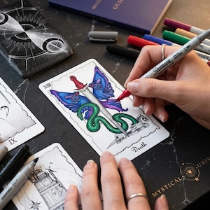 May include: A tarot card titled Death featuring a central sword, blue butterfly wings, and a green snake coiled around a skull. The card is being colored with a red marker. Other tarot cards, markers, and a book titled Mystical Universe are visible.
