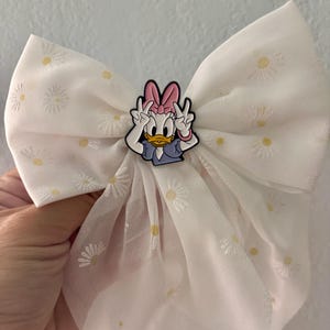 May include: A white hair bow made of sheer fabric featuring an embroidered daisy pattern with yellow centers. Centered on the bow is a character charm of Daisy Duck wearing a pink bow, making a peace sign with both hands, and wearing a blue top.