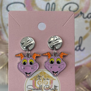 May include: Purple dragon face dangle earrings with silver metal tops. The earrings are on a pink card with the text "It's a Small Craft World".