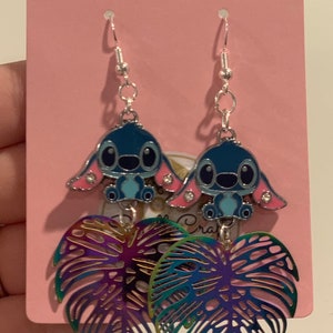 Peut inclure: Boucles d'oreilles pendantes avec un personnage de dessin anim&eacute; bleu et des oreilles roses, associ&eacute;es &agrave; des breloques en forme de feuille iris&eacute;e. Les boucles d'oreilles sont suspendues &agrave; des crochets d'oreille argent&eacute;s et pr&eacute;sent&eacute;es sur une carte rose.