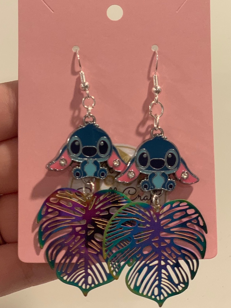 Peut inclure: Boucles d'oreilles pendantes avec un personnage de dessin anim&eacute; bleu et des oreilles roses, associ&eacute;es &agrave; des breloques en forme de feuille iris&eacute;e. Les boucles d'oreilles sont suspendues &agrave; des crochets d'oreille argent&eacute;s et pr&eacute;sent&eacute;es sur une carte rose.