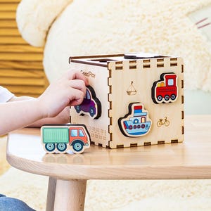 May include: A wooden shape sorter cube with vehicle cutouts, including a car, truck, boat, train, and bicycle. The cube is natural wood-colored, and the vehicles are painted in various colors. A child is playing with the toy.