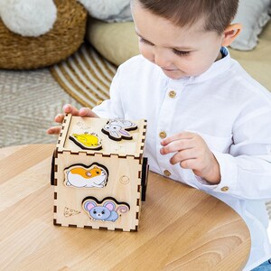 May include: A wooden animal puzzle cube with colorful animal shapes. The cube is made of light-colored wood and features images of a rabbit, a hamster, a mouse, and a yellow animal. A child is playing with the puzzle.