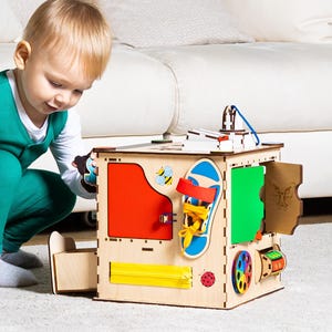 May include: A child plays with a wooden activity cube with various textures and activities. The cube has a red, yellow, green, and blue side with a butterfly, a zipper, a wheel, and a latch.