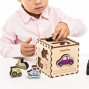 May include: A child plays with a wooden toy box with cutouts of different shapes. The box has the words "GoodPlay" printed on the side. The child is placing a wooden toy car into the box.