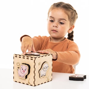 May include: A child plays with a wooden toy cube with animal faces. The cube has a pig, a sheep, and other animal faces. The child is placing a wooden animal piece into the cube.
