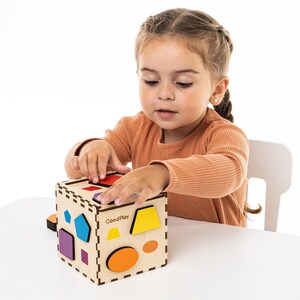 May include: A wooden shape sorter cube with various colored geometric shapes. The cube has cutouts for each shape and the text "GoodPlay" printed on the side. A child is interacting with the toy.