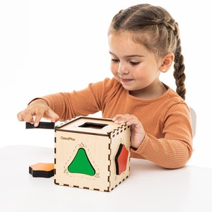 May include: A child plays with a wooden shape sorting box. The box has a green triangle, a red pentagon, and an orange hexagon. The box is labeled "GoodPlay".
