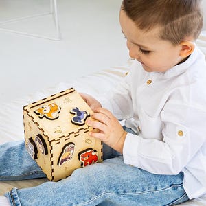 May include: A wooden cube puzzle with animal shapes. The cube is light brown with colorful animal cutouts on each side. The child is wearing a white shirt and blue jeans. The cube is approximately 5 inches in height.
