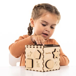 May include: A child plays with a wooden shape sorter toy. The toy has a variety of shapes, including a star, a circle, a square, and a pentagon. The toy is made of natural wood and has a light brown finish. The toy is labeled "GoodPlay".
